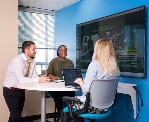 A group of people seating or standing around a table and tv monitor on the wall. 